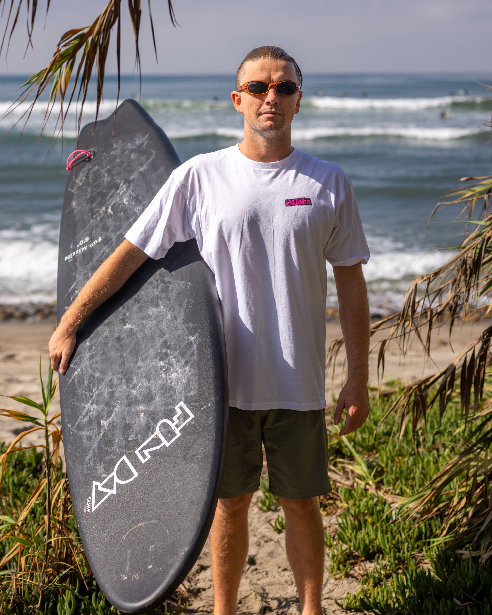 Fun Day Surf Aloha Tee in White worn with a surfboard at the beach, lifestyle view