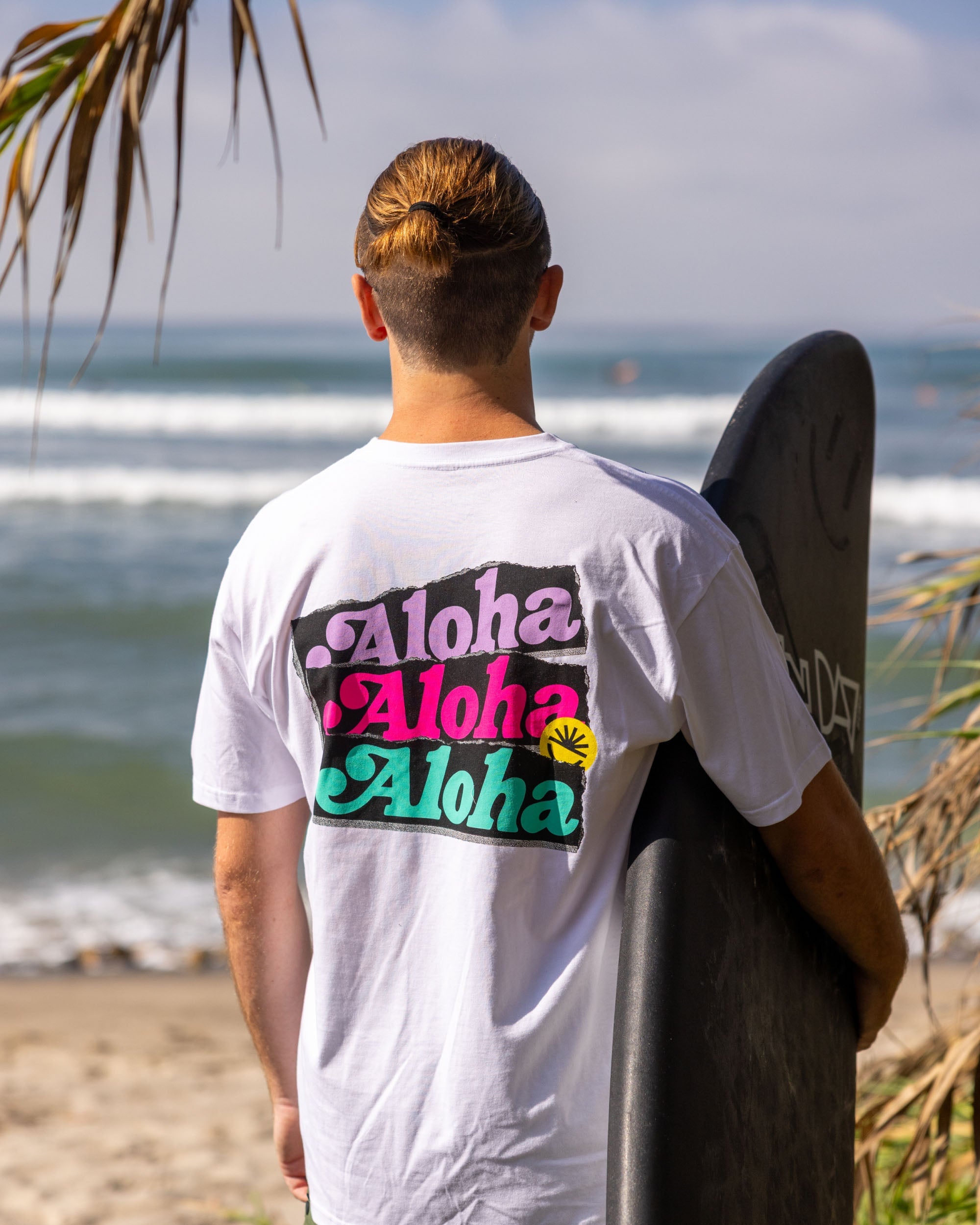 Fun Day Surf Aloha Tee in White back graphic worn with a surfboard at the beach, lifestyle view