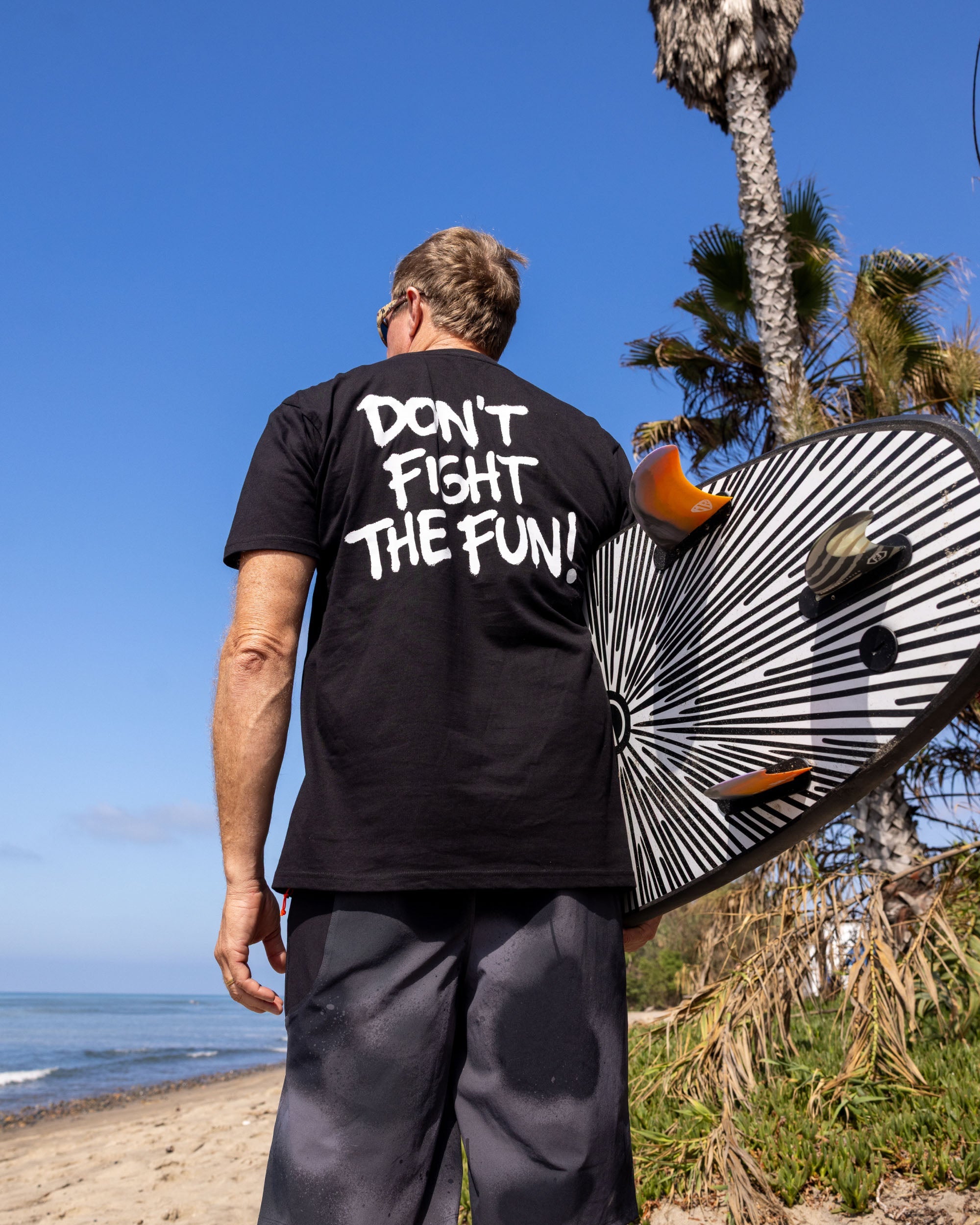 Fun Day Surf DFTF Tee in Black back graphic worn with a surfboard by the ocean, lifestyle view