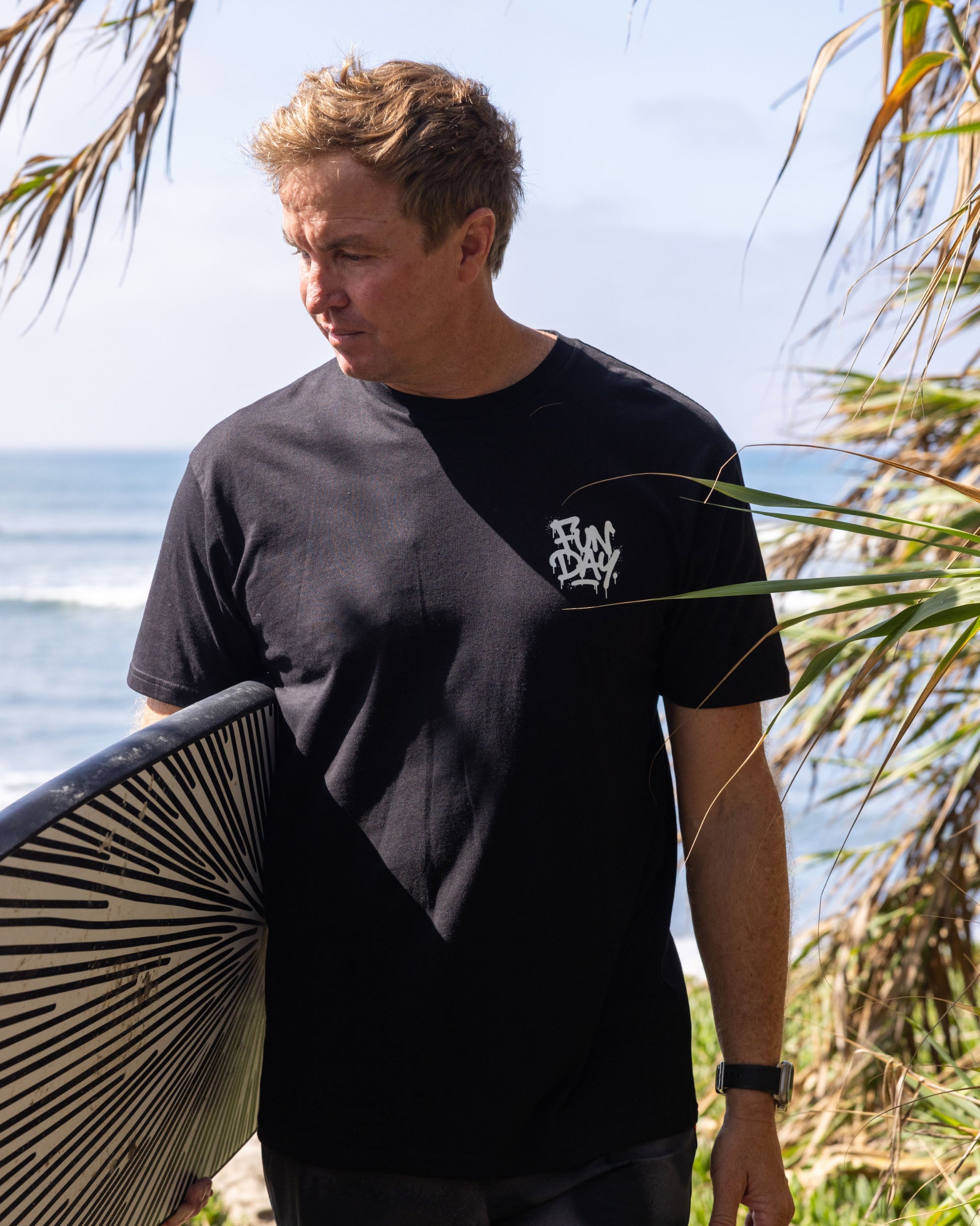 Fun Day Surf DFTF Tee in Black worn with a surfboard at the beach, lifestyle view