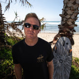 Fun Day Surf Framework Fill Tee in Black worn with a surfboard under the palms, lifestyle view