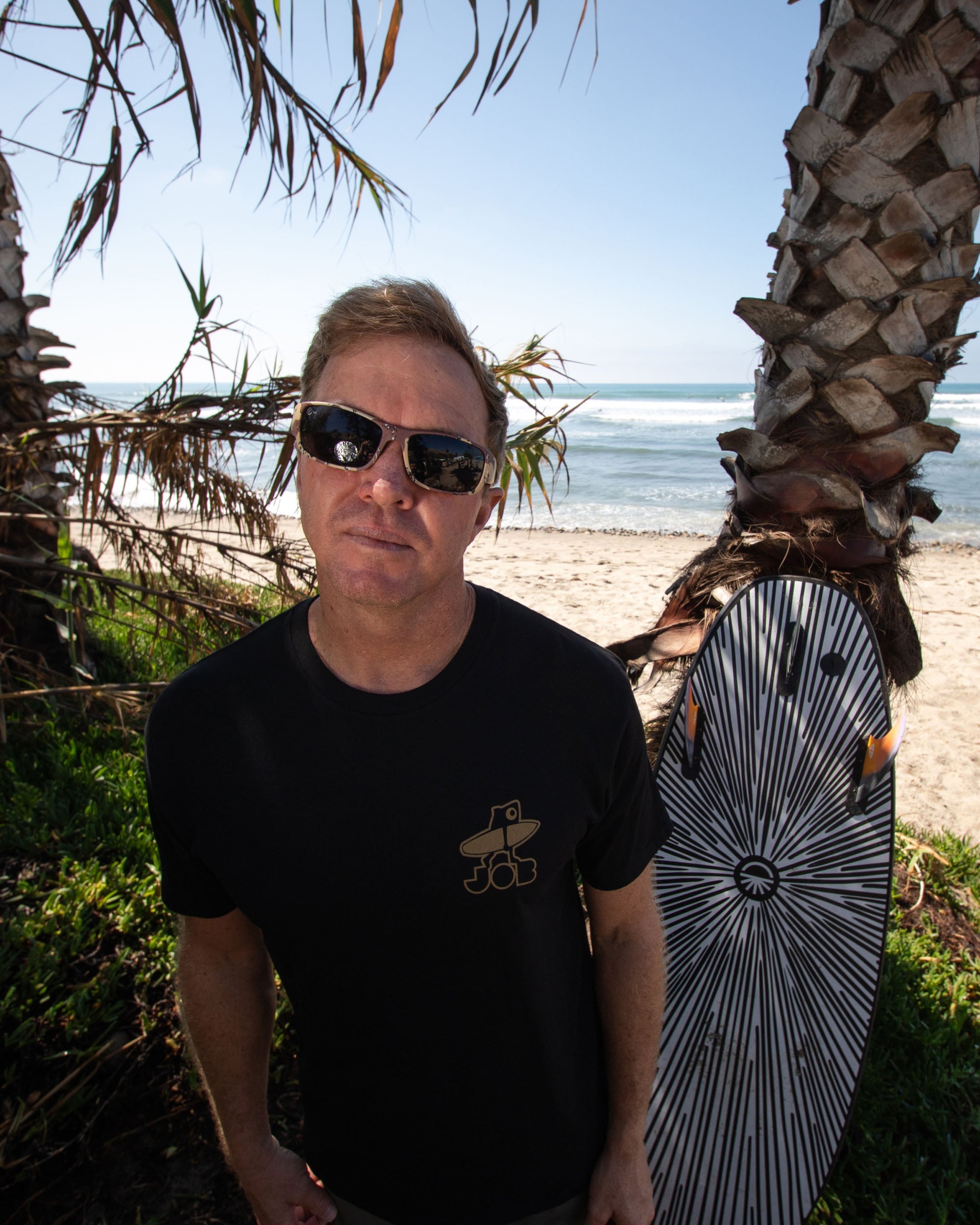 Fun Day Surf Framework Fill Tee in Black worn with a surfboard under the palms, lifestyle view