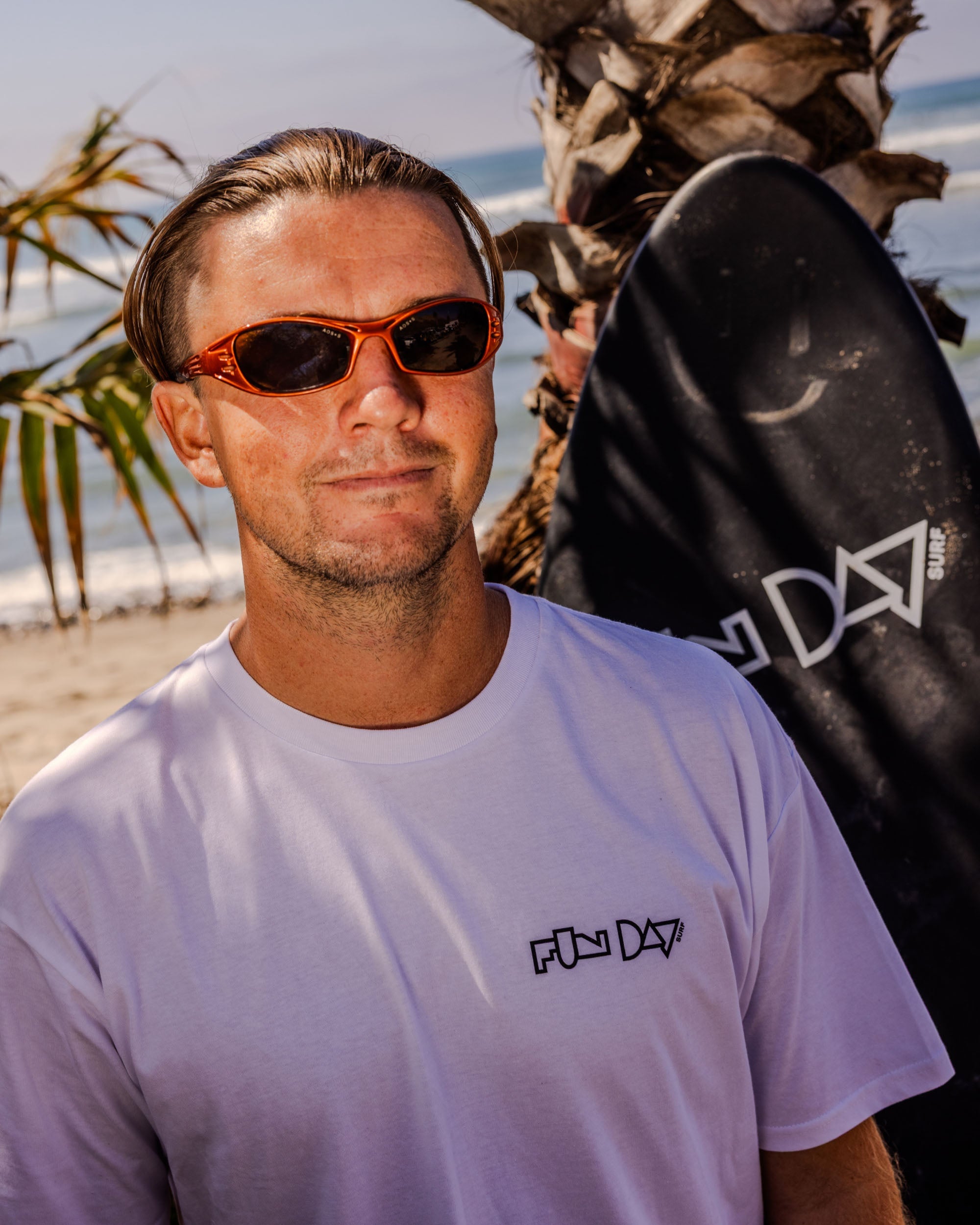 Fun Day Surf Framework Fill Tee in White worn with a surfboard at the beach, lifestyle view
