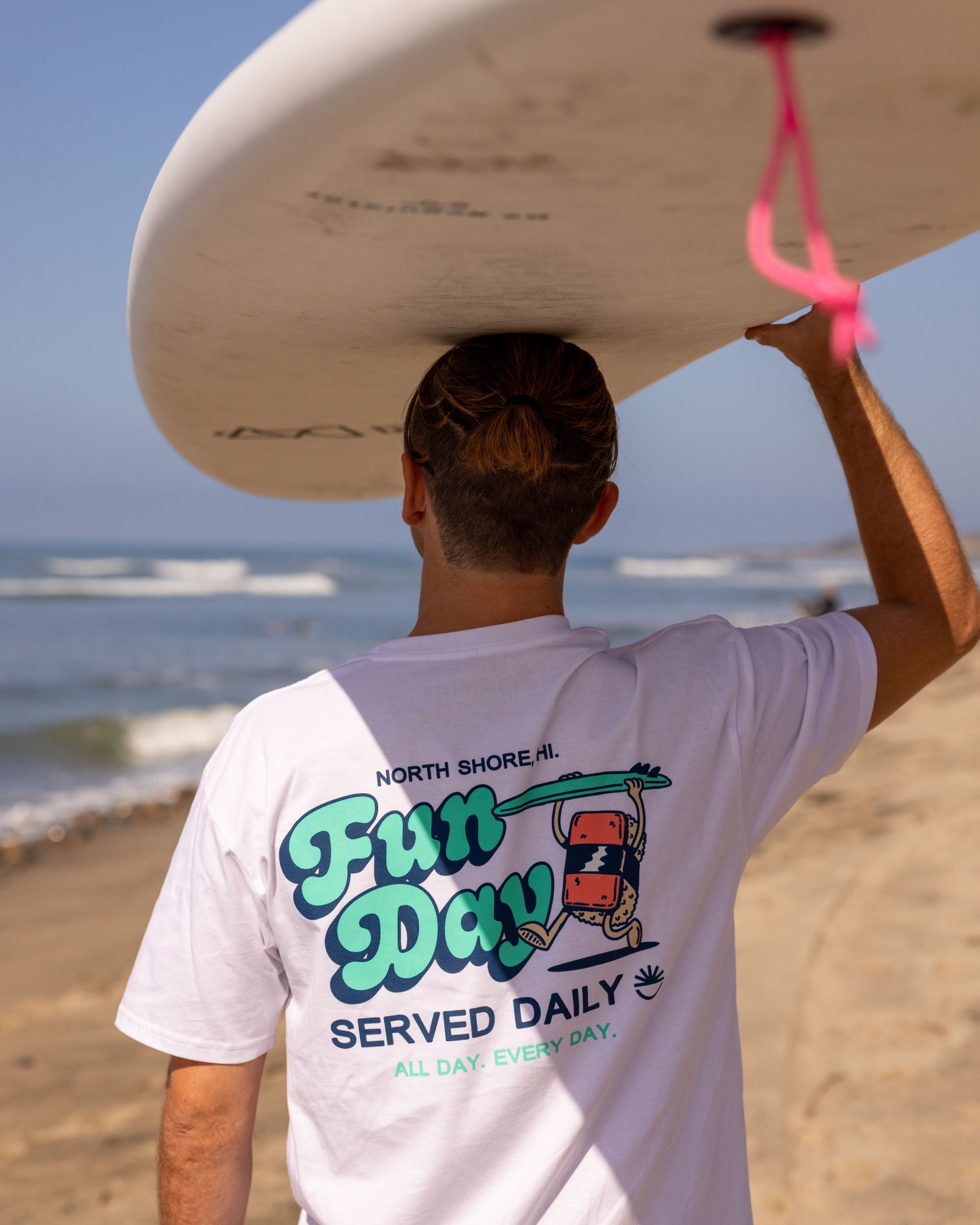 Fun Day Surf Musubi Tee in White back graphic worn carrying a surfboard, lifestyle view