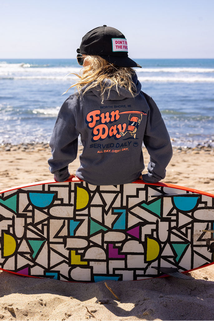 Fun Day Surf Musubi Youth Hoodie in Steel Blue back graphic worn on the beach with a surfboard, lifestyle view