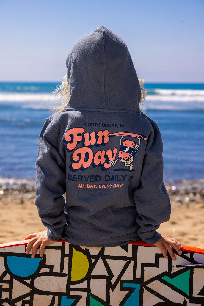 Fun Day Surf Musubi Youth Hoodie in Steel Blue back graphic worn holding a surfboard at the beach, lifestyle view