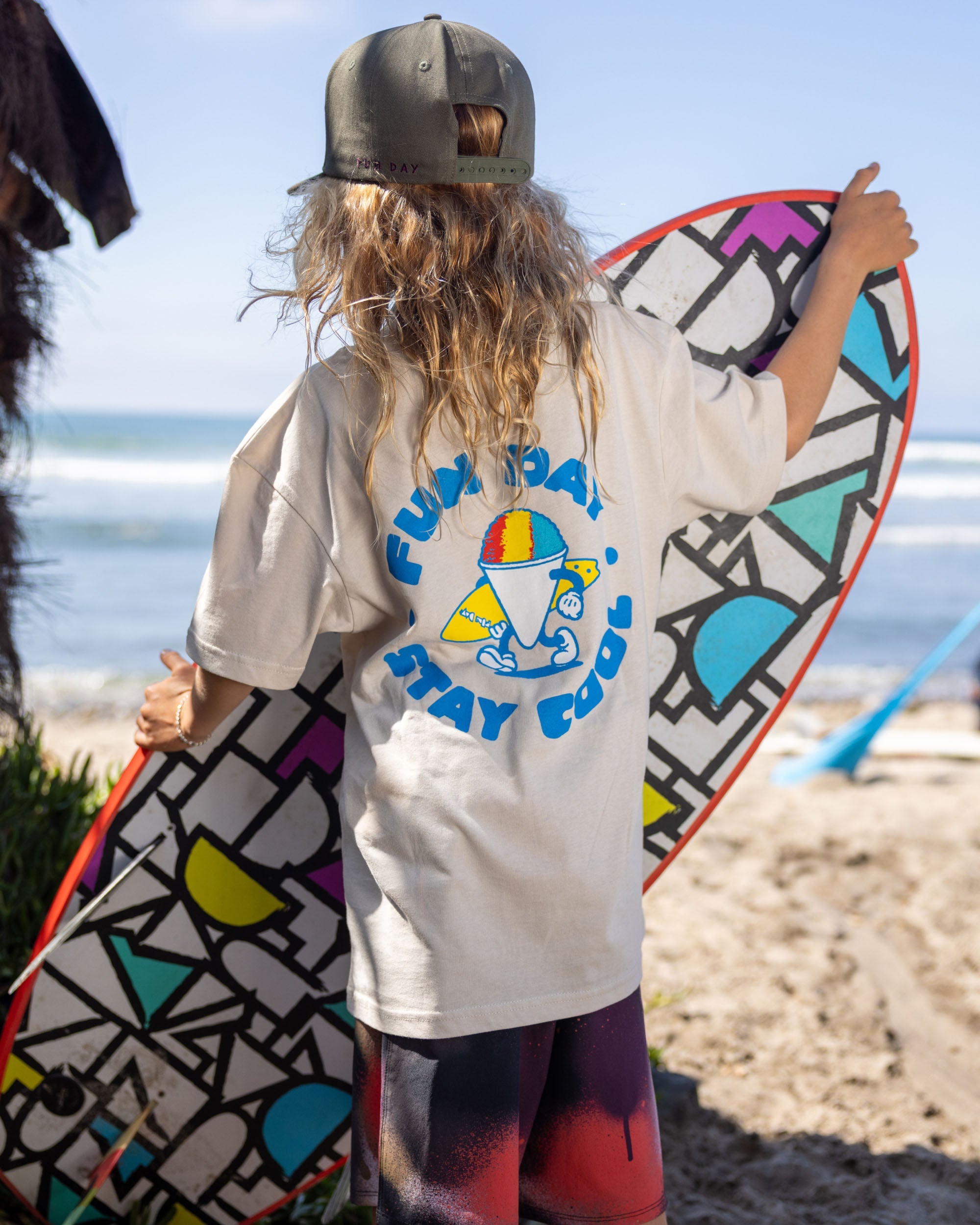 Fun Day Surf Stay Cool Youth Tee in Bone back graphic worn with a surfboard, lifestyle view